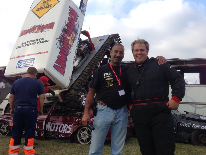 A group of people standing next to a truck - Pistonheads - The image shows two men standing next to a race car described as "Dead on the Track". They are both smiling and appear to be happy, possibly celebrating an event or cheering on companions. Behind them, a towering is visible, topped with a white vehicle. The men are dressed in casual clothing with red shirts. The backdrop of the image is filled with a cloudy sky.