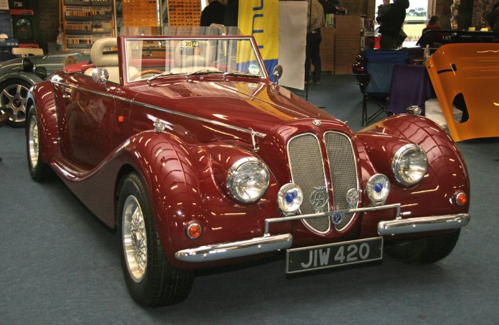 A red car with a red and white design - Pistonheads - The image shows a vintage car on display inside what appears to be an exhibition hall. The vehicle is a convertible with the top down, revealing its interior. It has a red and cream exterior color scheme and features distinctive design elements typical of classic cars from that era. The car is parked facing forward, allowing for clear viewing of both the front and rear aspects. In the background, there are other vintage vehicles visible, indicating that this might be a special event or exhibition dedicated to antique and classic automobiles.