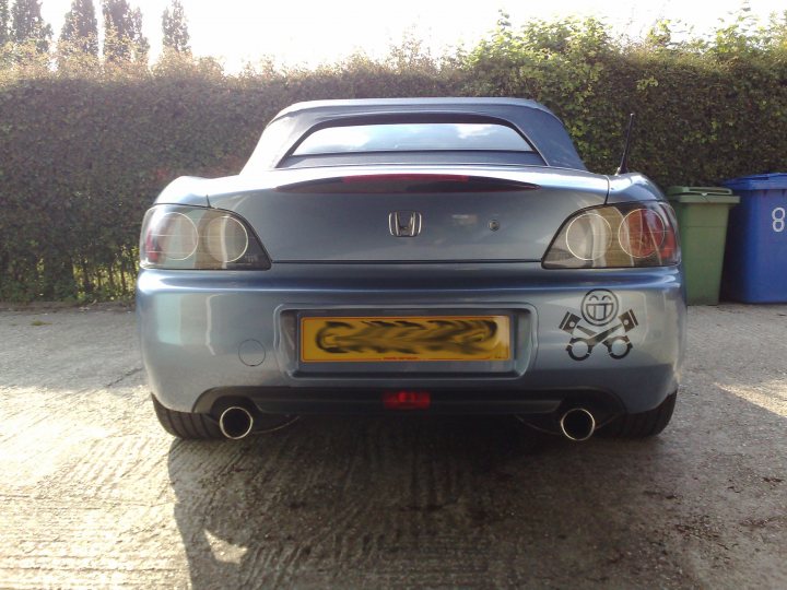 Pistonheads - The image features a rear view of a silver Honda sports car, parked on a concrete surface. It has a distinctive black and white plate with Japanese characters. The backdrop includes green plants and two large trash cans. The sun is shining on the car, casting a soft light and creating shadows on the ground.