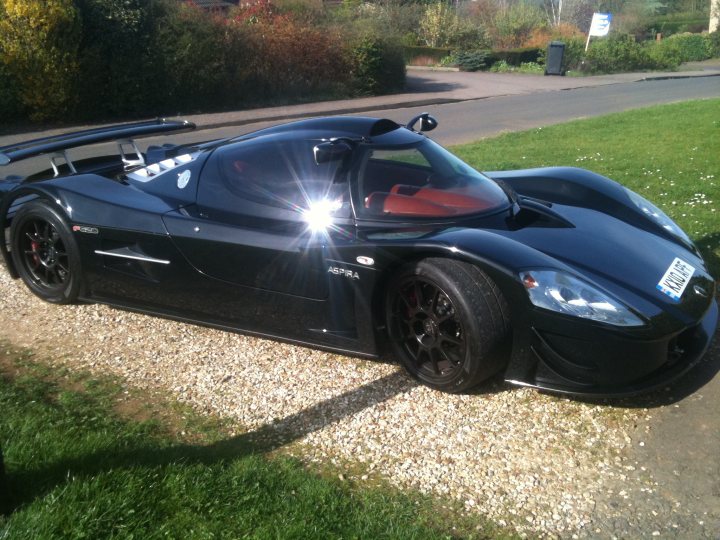 Pistonheads Spotted Northants - The image shows a unique black sports car parked on a gravel surface. The car appears to be a convertible with a sleek design and a large rear wing spoiler. It features butterfly doors which open outward, allowing access to the interior. The license plate of the car indicates it is registered in the UK. There is a grassy area to the right of the car, and in the background, sparse vegetation and a street light can be seen. The sky is overcast, suggesting a cloudy day.