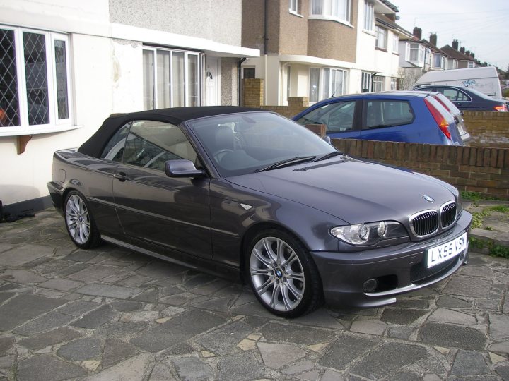 Sport Pistonheads - The image features a black BMW convertible parked in front of a house. The car appears to be in good condition, with a neatly closed soft-top roof. The driveway is made of cobblestones, providing a rustic charm to the scene. In the background, there are other vehicles and houses, suggesting a suburban setting. The visibility of the sky indicates that the photo was taken during daytime.