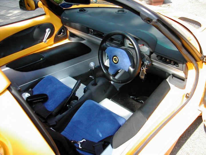 Sport 160 owners - Page 1 - Elise/Exige/Europa/340R - PistonHeads - The image displays the interior of a car, focusing on the front passenger side with the seat reclined. The seat is upholstered in blue, as are the side door panels and the floor mats. A steering wheel is visible in front of the seat, indicating that the driver's side is adjacent. The dashboard shows various control dials and a side-view mirror, and there are sun visors with reading lamps attached. The car's design suggests it might be from a luxury brand. There are no visible texts or distinctive markings in this close-up view.