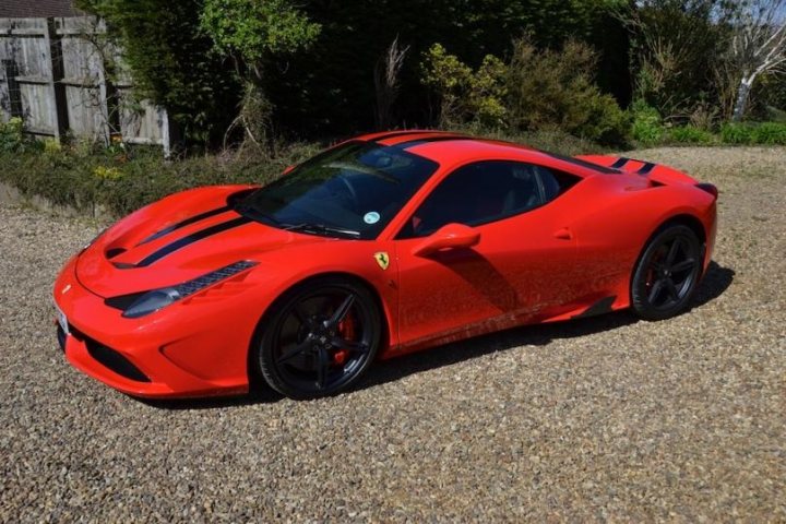 458 Speciale - Page 1 - East Anglia - PistonHeads - The image showcases a vibrant red Ferrari sports car parked on a gravel driveway. The car is facing to the left side of the image, showcasing its long curve of the hood and the elongated front arches. The black wheels with red Ferrari emblems and black tires contrast with the body. The backdrop includes overgrown vegetation suggesting a natural environment, indicating the car is likely in a residential or semi-rural area.