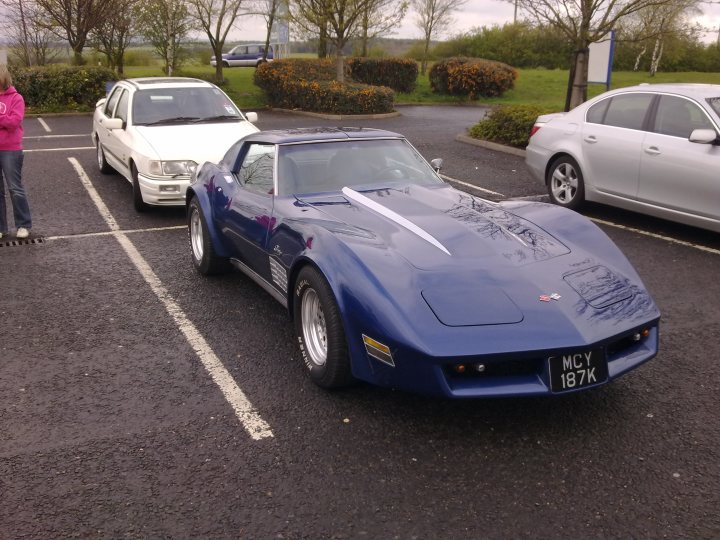 en route to hartlepool marina on drive it day - Page 1 - Corvettes - PistonHeads - This image shows a clear daytime scene in a parking lot. A vibrant blue convertible sports car is parked facing the camera, with the top down. Its sleek design and shiny paint add a touch of luxury to the setting. Behind the sports car, there is a white sedan parked at an angle. In the background, a few other vehicles are visible, and there are green bushes near the area. There is a person standing near the white sedan, adding a human element to the scene. The parking lot seems to be in a well-maintained area, as there are delineated parking spaces and clear road markings.