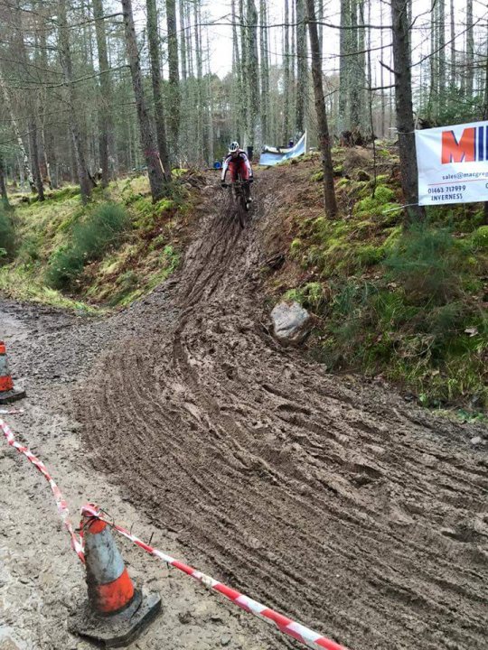 The "Photos From Today's Ride" thread... - Page 233 - Pedal Powered - PistonHeads - The image captures a dramatic scene on a dirt trail cut through a forest. At the center of the frame, a cyclist, clad in a helmet and backed by a white jersey, is halfway through a dizzying jump on their bicycle. The bike seems to be mid-air, suggesting significant speed or momentum.

The path is temporarily closed due to possible damage; a red rope and several orange cones have been laid out to warn potential passersby. The dirt itself appears to be uneven, contributing to the overall ruggedness of the scene.

The backdrop is filled with trees, some taller than the cyclist, establishing a natural and wild setting. A large white sign, partially obscured by an orange cone, is also visible, though the text on it is not clear. The entire image conveys a sense of adventure mixed with caution.