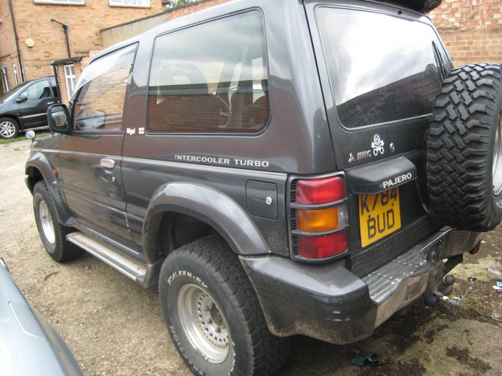 Offroader Cheap Pistonheads - The image depicts the rear view of a black Jeep SUV parked on a dirt driveway. The vehicle features a spare tire mounted on the back, and a red tail light that matches the color of the license plate. A license plate at the back reads "K84 PAJERO." The Jeep has side steps and a black bumper. The setting suggests an outdoor location, possibly a residential area, as a brick building is partially visible in the background.