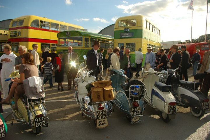 Revival - First Time and Excited - Page 1 - Goodwood Events - PistonHeads - In the bright daylight, a lively event is unfolding on a city street where people are standing around, chatting, and admiring the collection of vintage transit buses and scooters. A large double-decker bus, painted in vibrant yellow, green, and red, is prominently parked with its doors open, revealing an old-fashioned interior. Beside it, a single-deck green bus adds to the vintage charm of the scene. Parked in front and on the side of these buses are two white motor scooters, one prominently displaying an English license plate with the letters "A10" clearly visible. The street itself is lined with buildings, and a lone flagpole stands in the background, adding a touch of patriotism to the urban setting.