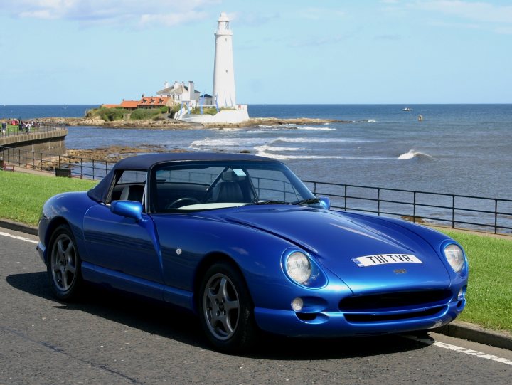 Wow! A sunny day... - Page 1 - North East - PistonHeads - The image presents a serene coastal scene. A sleek, bright blue contemporary sports car is parked on the road, which is adjacent to a grassy area. The car is facing the viewer, allowing a clear view of its design. A lighthouse, a white brick structure with a pointed top, stands in the background, adding to the coastal charm. In the sea nearby, there are seagulls, visible as small white figures against the vast expanse of the ocean. The sky is clear and blue, suggesting a sunny day. The overall atmosphere of the image is calm and relaxed.