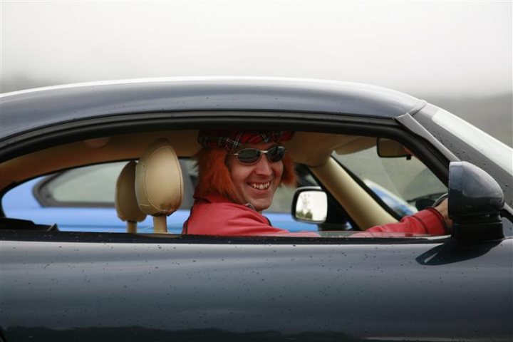 Pistonheads - In the image, there's a man with reddish-orange colored hair, also known as ginger hair, who appears to be very happy as he is smiling brightly. This man, wearing sunglasses and a scarf, is seated in a convertible sports car, the top of which is down. The car has two distinct doors or sides that the man is occupying. The car's interior is visible, suggesting the seat is a tan or beige color. The car is seen from the perspective of the open door, with the man's friends, notable for their colorful headbands, captured in the side mirror of the vehicle. The sky forms the backdrop of the image, indicating that the photo might have been taken during the day.