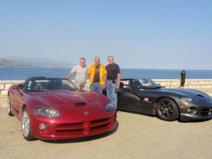 Celebrating 20 years of ... - Page 1 - Vipers - PistonHeads - The image captures a scenic view of the ocean, where three men are standing next to two sports cars parked by the water. The men are dressed casually, with two of them sporting jackets and the third in a t-shirt. The cars, one glossy red and the other a sleek black, are parked on a paved area near the edge of a platform, which overlooks the vast expanse of the sea. Above them, the sky appears to be a clear blue, suggesting a bright and sunny day. The gentle lapping of the waves against the rocks on the water's edge adds to the serene ambiance of the scene.