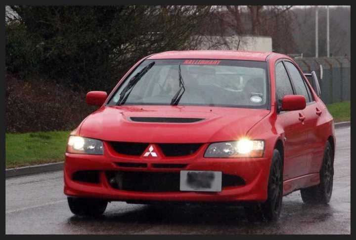 Post your Evo pics - Page 4 - Jap Chat - PistonHeads - The image shows a red compact vehicle traveling on a wet road where rain has left the surfaces shiny. The car is captured in motion, and the reflections from the water suggest it might have recently rained or be raining at the time the photo was taken. The vehicle has a prominent grille with the Mitsubishi logo and features a set of fog lights integrated into its design. The license plate is not visible for privacy reasons. Due to the motion blur and the wet surface conditions, the details of the surroundings are indistinct.