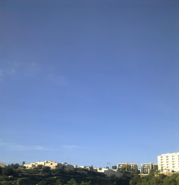 Pistonheads Work Drive - The image shows a bright, clear blue sky overhead expansive green hillsides. In the distance, there are modern apartment buildings with several floors, indicating a residential area. At the foot of the hills, partial tree cover is visible, providing some green contrast to the concrete structures. The lower part of the image shows what appears to be the outline of a city, based on the architecture and the terrain.