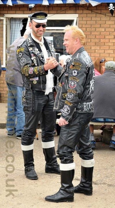 Jack's Hill Ton Up Day - Page 1 - Biker Banter - PistonHeads - The image captures a moment on a street, where two individuals, both dressed in leather jackets, black pants, and boots, are the focal point. They appear to be the members of a motorcycle gang or a similar group, as indicated by their matching attire and symbols. The individual on the right seems to be actively engaging with the one on the left, perhaps in a friendly manner, while the other individuals in the background contribute to the lively street scene. The overall atmosphere suggests a sense of camaraderie and shared identity among the two dominant figures.