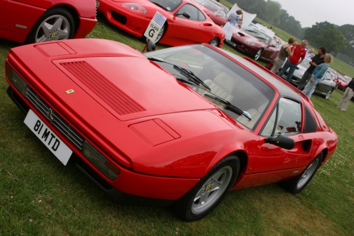 F353 TRY - Anyone know it? (328 GTS) - Page 1 - Ferrari V8 - PistonHeads - The image features a vintage red sports car parked on a grass field with a group of people standing around. The car has Italian-made specifications listed on the front, indicating its origin or racing history. The vehicle's speedometer is prominently visible in the foreground, extending over the vehicle's front wheel. The surrounding area appears to be an outdoor event or open-air private area where several other cars are parked behind the red sports car, implying a gathering or display of vehicles.