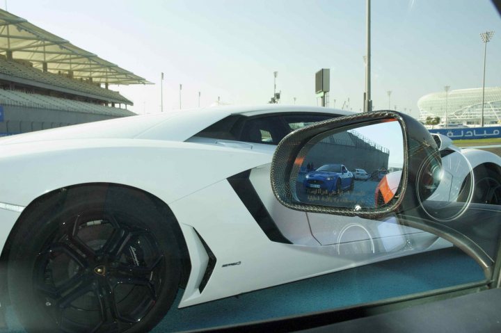 Pistonheads - The image captures a modern, white sports car reflected in a rear-view mirror. The car is parked on a track with bright daylight. In the mirror's reflection, multiple buildings are visible, suggesting the location is near a sports arena. The spokes of a bicycle tire can be seen in the foreground, adding depth to the image. The car's sleek design and the architectural style of the buildings hint at a contemporary setting.