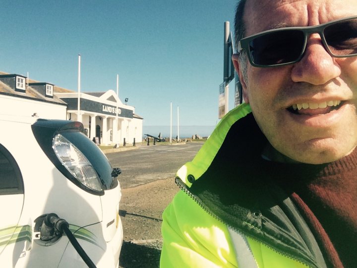 Lands End to John O'Groats - Page 1 - EV and Alternative Fuels - PistonHeads - The image features a man dressed in a yellow safety vest standing beside a white car. The man appears to be in mid-laughter, creating a joyful atmosphere. The car has an environmentally-friendly topping, and there is a small building with a sign that reads "Land's End" in the background. The man is wearing sunglasses, and the weather seems to be clear and sunny.