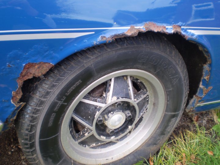 Scrap the Stagg, or give it a new home? - Page 1 - Triumph - PistonHeads - The image showcases a large tire attached to a bright blue vehicle. The tire displays a pattern of text that reads "B XXX S." The rim of the tire is silver, contrasting with the blue exterior of the vehicle. Interestingly, the vehicle does not appear to have a fender around the wheel, and there is evidence of rust on the metal surfaces of the car. The scene is set on a grassy surface, suggesting that the vehicle may be parked or abandoned outdoors.