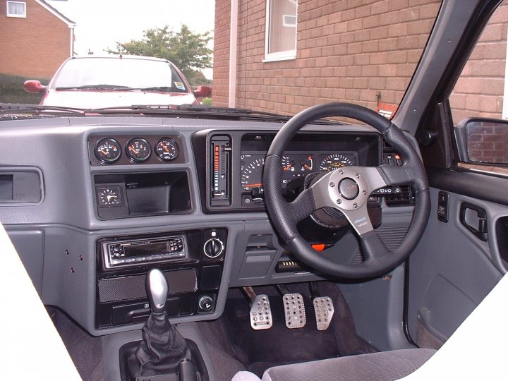 My Sierra XR4x4 Turbo Technics - Page 1 - Readers' Cars - PistonHeads - The image shows the interior of a vehicle, focusing on a steering wheel. The steering wheel is at the center of the dashboard, featuring a variety of buttons and gauges. There is a gear shift visible to the right of the steering wheel. The background reveals a brick building and cars, suggesting the vehicle is parked in a residential or urban area near the road. The image appears to be taken through an open car door, providing a view into the vehicle from the passenger side.