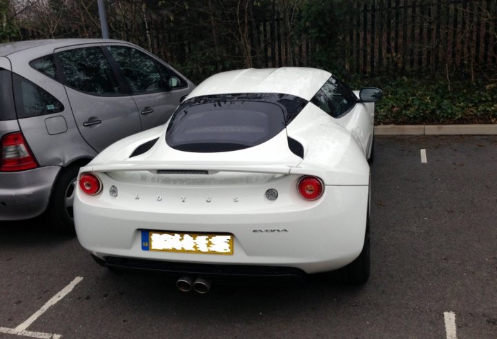 Purchased Larini + De-cat - Page 1 - Evora - PistonHeads - The image shows a parking lot with a white Lotus car parked next to a grey hatchback. The Lotus car has a black, retractable roof and is designed with aerodynamic lines. It's positioned in the middle of the parking space, and the rear of the car is documented by a yellow license plate. The car's design and color contrast with the more utilitarian-looking grey car. Both vehicles are parked on an asphalt surface, with the car in focus and the background featuring slatted fencing and a few trees.