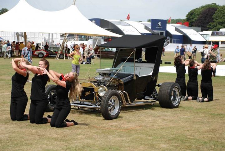 Goodwood Pistonheads - The image captures an outdoor scene at an auto show or similar event. A group of young women, possibly dancers, are staging a performance or demonstration with an old-fashioned vehicle, which appears to be a drag car given its elongated chassis. The women are dressed in matching black attire, and they are kneeling down around the car to simulate spiritual veneration or a stylized performance. The setting is surrounded by spectators, and there are advertisements and a tent in the background. The atmosphere suggests an entertaining and theatrical occasion.