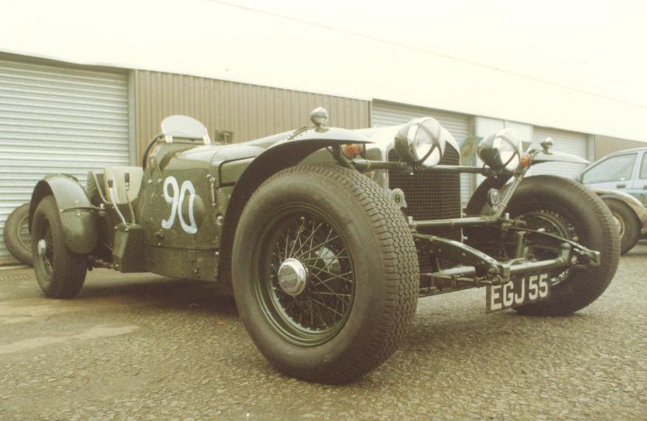 Egj Pistonheads Bentley Derby Special - The image shows a vintage race car parked in a lot next to a modern truck. The car, numbered 9 0 5, has a streamlined body painted in a classic racing livery of red and white. It has large silver headlamps and black spoked wheels, characteristic of its era. The vehicle displays signs of use, with visible scratches on the body. The setting appears to be an industrial or commercial area, indicated by the buildings in the background.