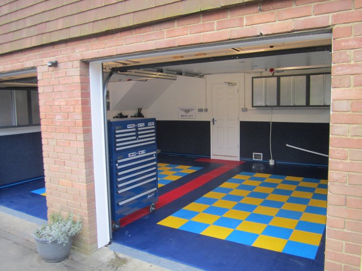 Who has the best Garage on Pistonheads???? - Page 189 - General Gassing - PistonHeads - The image shows an indoor space that appears to be a garage or workshop within an alleyway of a residential area. A large door, likely used for driving or accessing the interior, is open, providing a view of the room. The interior of the space has a tiled floor with alternating squares of blue and yellow color, reminiscent of a checkerboard pattern, that covers the flooring area. On the left side, there is a blue tool shelf which is likely filled with various tools and equipment not fully visible in the image. The garage wall features a white door and a black panel with some text on it; however, the text is not legible in the image. Additionally, a single plant is placed on the left side of the image, adding a touch of greenery to the otherwise industrial space.