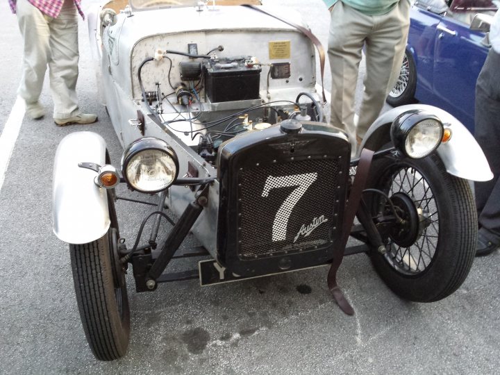 COOL CLASSIC CAR SPOTTERS POST!!! Vol 2 - Page 62 - Classic Cars and Yesterday's Heroes - PistonHeads - The image shows a vintage race car from the early 20th century parked in an asphalt lot, likely an event or exhibition of classic cars. The car appears to be under restoration, judging by the visible parts under different colors of paint. An identifiable feature is the number "7" on the side, suggesting it may be a historical or special edition vehicle. In the background, a person in a green top seems to be observing the car, along with a few more individuals, creating a casual yet attentive atmosphere at the car show.