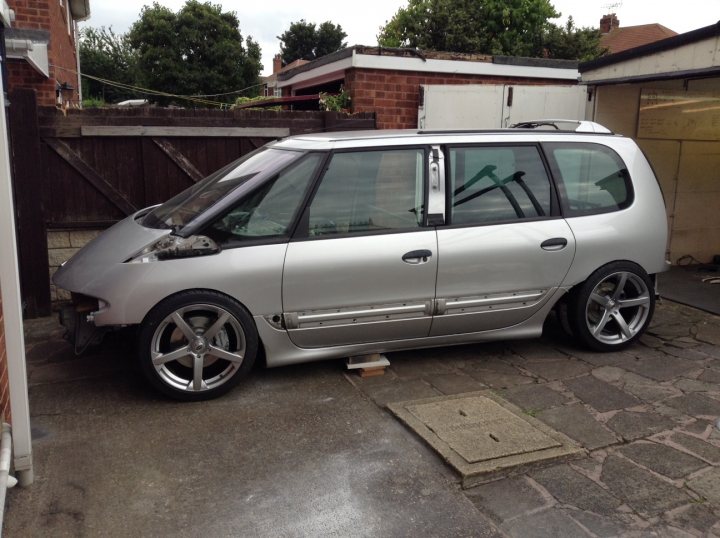 Lexus V8 with NOS in a Renault Espace - yeah lets do it !  - Page 12 - Readers' Cars - PistonHeads - The image depicts a grey, van-style car parked near a brick wall, close to a shed. The car's design includes a large front window and a noticeable wheel size that could suggest a sportier or aftermarket modification. The body of the car appears to be made of metal, as indicated by the shiny surface and presence of a large dent on the side. The setting suggests a residential area with a private driveway.