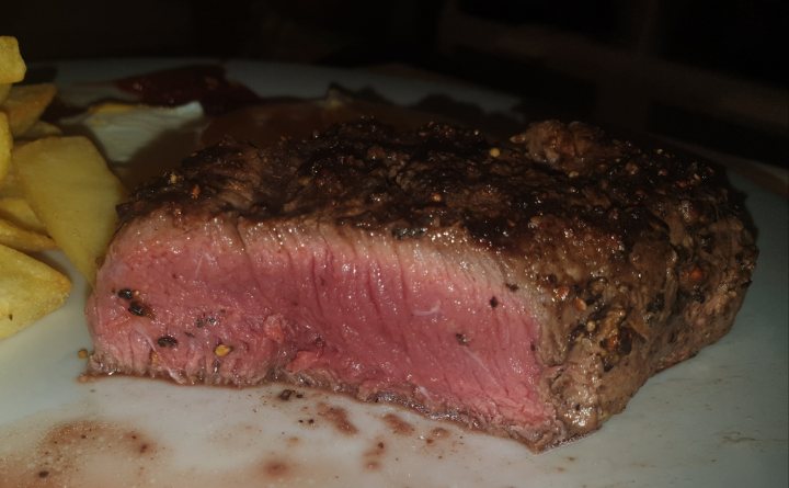 THE  STEAK THREAD, served a la Man. - Page 55 - Food, Drink & Restaurants - PistonHeads - The image features a large, raw steak with a rich color, prominently placed on a white plate. The steak appears to be well-marbled, suggesting high quality. To the right of the plate is a side of fries, crispy and golden brown. Behind the main dish, there's a small amount of red sauce or ketchup. In the upper left corner of the image, there is a blurred figure that seems to be holding or preparing something, although their face isn't clearly visible. The overall scene suggests a meal preparation or presentation.