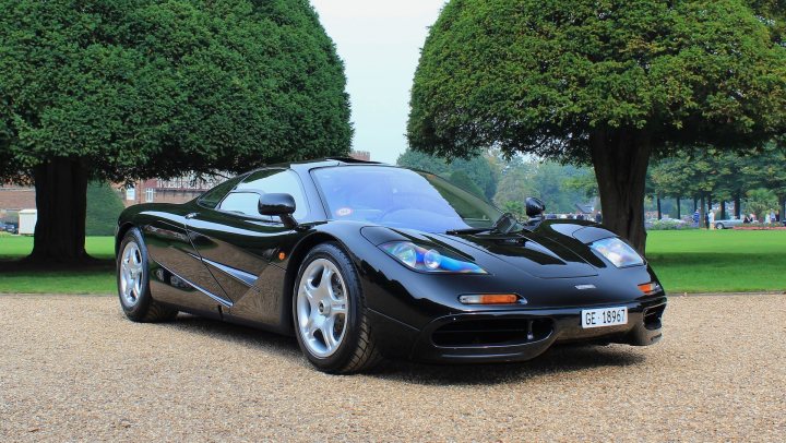 A black and white photo of a classic car - Pistonheads - The image features an elegant black sports car parked on a gravel road. The car is positioned in front of a pair of trees with green, rounded canopies that appear healthy and well-maintained. The vehicle's sleek design, with its long and low hood, aerodynamic curves, and large round LED headlights, gives it a distinctive and sporty appearance. In the background, there is a glimpse of people, suggesting a public place or park where the car is on display.