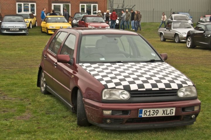 Pistonheads Bye Accordian Golf - The image portrays a gathering in a parking lot where a multitude of cars, both vintage and modern, have been meticulously parked. The focus of the image is a distinctive checkered car with a license plate that reads "R139 XLG". This car has a black and white checkered hood, adding a touch of boldness to its overall appearance.

The event seems to be well-attended, as evidenced by the numerous spectators scattered throughout the lot, all seemingly absorbed in admiring the array of cars on display. The backdrop of the parking lot provides a contrast to the colorful diversity of the vehicles.

The checkered car is situated such that it stands out from the rest of the cars, drawing the viewer's attention to its unique design. Its position in the middle of the frame contributes to its prominence. The other cars, parked neatly, add depth to the scene, demonstrating the vastness of the parking lot.

Overall, the image captures a lively car show, with the checkered car as the star of the event.