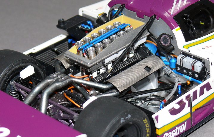 Tamiya 1/20th Jaguar XJR9-LM - Page 14 - Scale Models - PistonHeads - The image displays an intricate model of a race car with its engine partially exposed. The engine bay includes various components such as cylinder heads, connectors, and metallic parts with visible nuts and bolts. The car has vibrant purple and yellow racing colors with the word "SEGA" prominently displayed. The track or road surface where the model car is placed is not visible.