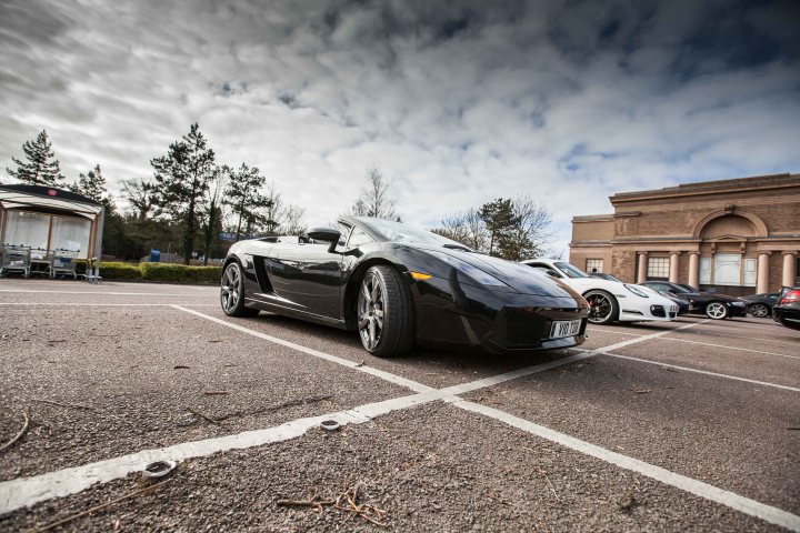 Supercar meet at Flying Horse PH clophill 22/3/15 - Page 9 - Herts, Beds, Bucks & Cambs - PistonHeads - The image features a black, convertible sports car parked on a lot. The car is sleek and shiny, likely a convertible or luxury model, and it appears to be the central focus of the image. The surrounding environment includes a partial view of other vehicles and a building in the distance. The lot is marked by white lines, and there are other cars parked in the vicinity. The darkened mood of the image, with its overcast sky and muted colors, suggests perhaps an evening or cloudy day setting.