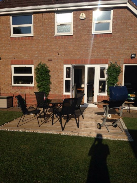 Question Regarding Moving Soil Pipe For Extension??? - Page 1 - Homes, Gardens and DIY - PistonHeads - This image captures a serene outdoor setting in front of a brick house. A wooden deck, neatly bordered by a white border and shaped like a large L, serves as the focal point. It's furnished with a black patio set, complete with a table and four chairs, inviting one to relax and enjoy the space. The house, painted in a soothing light brown color, stands as an elegant backdrop, with a window symmetrically placed on the right side of the frame. Above the deck, the sky stretches out in a tranquil expanse, dotted with lush green trees that add a touch of nature to the scene.