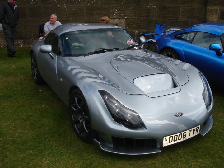 Attempt Pistonheads Chatsworth - The image captures an outdoor gathering of four people standing next to various types of cars. The most prominent vehicle is a sleek, silver sports car with striking black wheels. Its aerodynamic design and the shiny exterior suggest it's of high performance quality. The car is parked on a grass field, indicating a casual, possibly festive, setting.

In the background, two other cars can be seen. One is a vibrant blue convertible, and the other is a classic sports car in a rich shade of orange. The contrast between the oxidized red bare wall and the bright colors of the cars adds a sense of vibrancy to the scene. The positioning of the cars and people suggests a social event or a car show, where the vehicles are being admired or displayed.