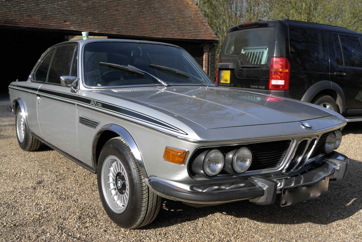 just bought a m3 CSL.  - Page 16 - M Power - PistonHeads - The image is a photograph of a silver BMW car, likely an older model, parked on a gravel surface. Behind the BMW is a modern black van. On the dashboard of the BMW, you can see two coffee cups with lids. The car and van have their respective head and tail lights illuminated. In the background, there is the sight of a building with a brown roof.