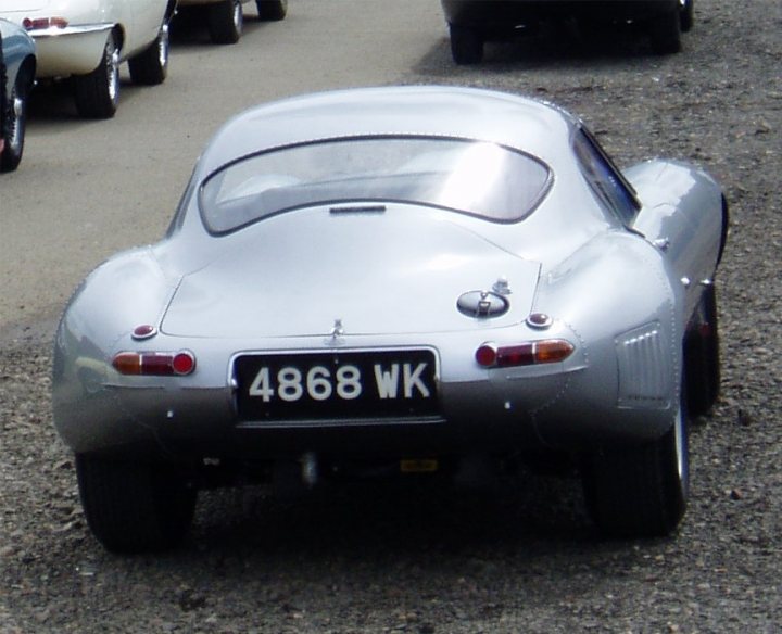 Pistonheads - The image shows a silver sports car parked on a gravel driveway. The car is positioned diagonally across the frame, with its right rear tire turned slightly to the left. The vehicle's license plate is white with black numerals and letters. The car's sleek design features a long hood, prominent rear spats, and rounded keel panel. The background reveals other cars parked nearby.