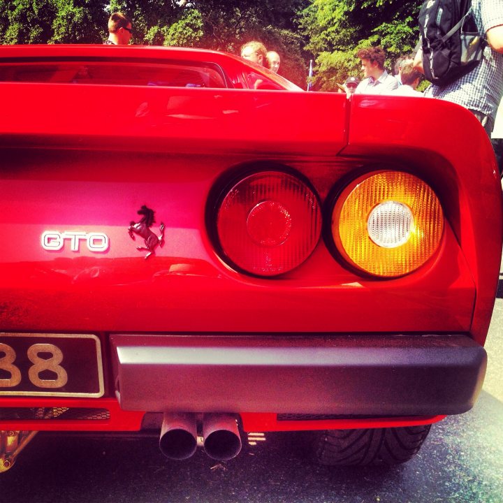 Post your Pictures here: Festival of speed 2013 - Page 1 - Goodwood Events - PistonHeads - The image features a striking red Ferrari, its sleek curved lines and the iconic logo on the back are unmistakable. It is parked, with its taillights prominently featured. The scene is set outdoors, under a clear sky, adding vibrancy to the car's glossy finish. In the background, there are people, suggesting a social gathering, but the focus remains on the captivating presence of the Ferrari.