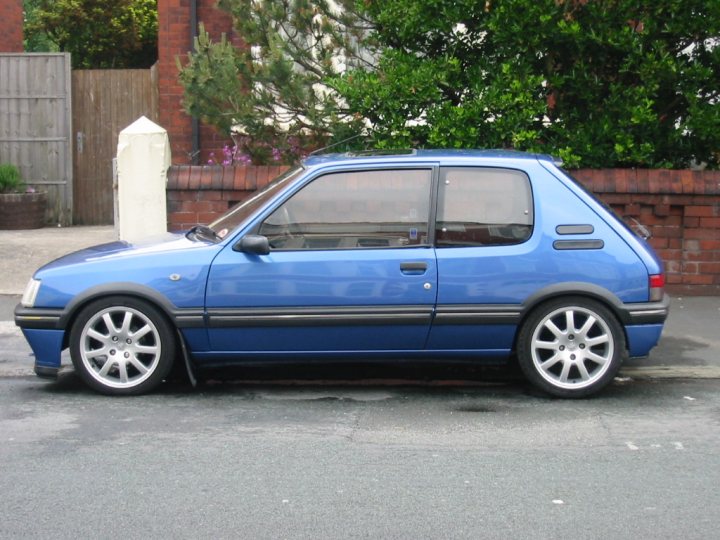 Pistonheads - The image displays a blue two-door car parked on the street. The vehicle's color scheme consists of a light blue upper half and a darker blue lower half, with grey stripes outlining the doors and a matching grey stripe running across the hood. The advertising code appears on the side. The car's wheels are black and complement the silver base. The side windows are down, revealing an empty interior. The car is positioned on a patch of asphalt with a brick building visible in the background. The setting suggests an urban residential area, with a metal fence, plants, and a hydrant on the sidewalks.