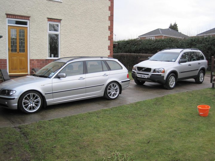 Types Pistonheads - This image depicts a suburban scene featuring two vehicles parked in front of a house. The first vehicle, a silver station wagon, has its front door wide open, revealing an interior space with the driver's seat visible. The second vehicle, a silver SUV, is parked directly behind the station wagon with its side profile facing the viewer. Both vehicles are parked on a concrete driveway that leads towards the house. On the right side of the image, there is a potted plant on the ground, and the vegetation around the house is noticeable. The sky appears to be overcast. The scene suggests a residential area with clear visibility of the vehicles and their immediate surroundings.