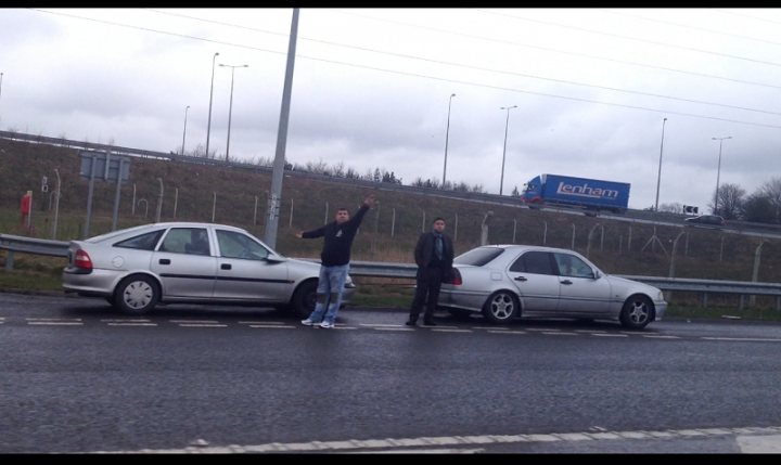 Odd Scam: Being flagged down on the Motorway! - Page 1 - General Gassing - PistonHeads - The image shows a group of three individuals standing on the side of a road. The person in the center is holding up his arms and appears to be mid-action, possibly dancing or gesturing. The two individuals on each side stand with their arms by their sides, looking towards him. They are all outdoors, indicated by the presence of a car parked on the side of the road and a truck visible in the background. The scene suggests a casual, informal gathering or event, possibly on a cloudy day, considering the somber overcast sky.