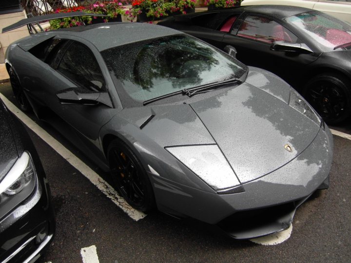 Vol Pistonheads Spotted - In the image, a sleek gray sports car is parked on the side of a parking lot. Its glossy exterior gleams under the sunlight, and the car is facing towards the left side of the image. The car is parked next to a row of black cars, making a noticeable contrast. The background features a variety of colorful flowers in pots, adding a touch of nature to the urban setting. A red and white building can also be seen, contributing to the overall composition of the scene.