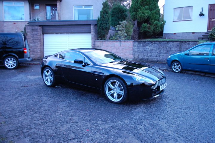 Pistonheads - The image captures a residential scene featuring a black Aston Martin sports car parked on the driveway of a house. The car, which stands out due to its sleek design and shiny finish, is positioned in front of a garage. The house itself is typical of residential architecture with a garage attached. Another blue car is parked nearby, possibly belonging to the house's occupants. The overall atmosphere suggests a quiet, suburban setting.