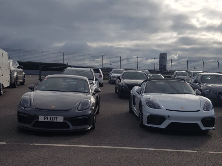 The new 718 Gt4/Spyder are here! - Page 65 - Boxster/Cayman - PistonHeads - The image showcases a parking lot filled with various sports cars. Two Ferrari models are prominently parked side by side, one of them is a convertible model. Other cars include a Lamborghini and a Porsche. The setting appears to be at an airport or airfield, as suggested by the presence of control towers in the background. The ground is covered with snow, indicating that the photo was taken during winter. There are also several people scattered around the area, possibly owners or enthusiasts admiring the vehicles.