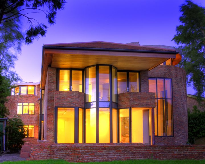 Double Aluminium Pistonheads Glazing - The image showcases a modern two-story house under the purple evening sky. The house is constructed with red brick and has a flat roof. The most striking feature is the presence of large, illuminated glass windows which bathe the interior with warm, yellow light, creating a contrast with the darkening sky. There are trees visible in the background, and the image is captured from a perspective slightly lower than the entrance of the house, emphasizing its architectural features.