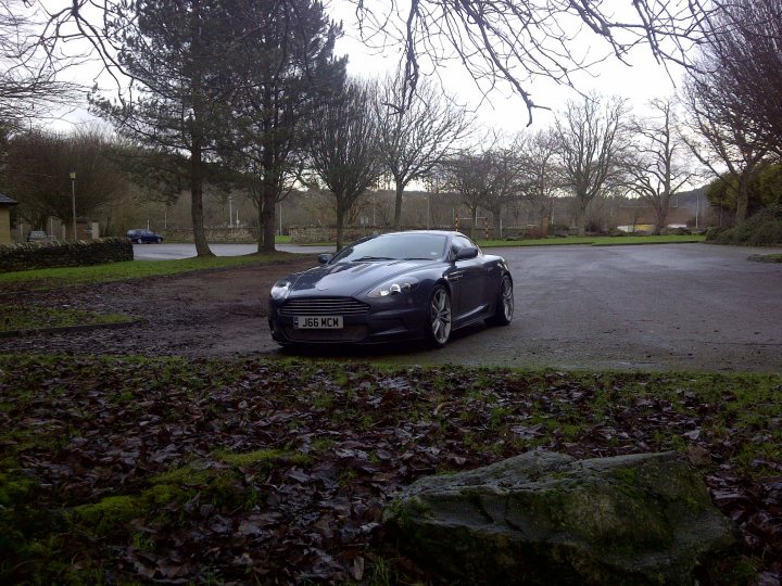 DBS wheels on DB9 - (more or less) - Page 1 - Aston Martin - PistonHeads - The image shows a stylish silver Aston Martin car parked on a wet pavement. It's an overcast day, and the car is partially shielded from the viewer by the foliage of trees in a grassy area. The vehicle is positioned parallel to the curb and appears to be an executive sedan, based on its sleek design and size. The surrounding landscape features natural elements such as trees and rocks, contributing to a serene and somewhat secluded setting.