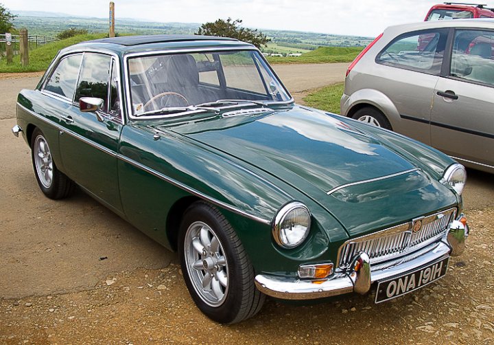 An old car is parked in a parking lot - Pistonheads - The image showcases a vibrant green vintage car parked on a gravel surface. It has two doors and a small rear window, reminiscent of classic designs. The car's round white headlights and silver-toned front grill, along with the spoke wheels, lend it a distinctive and nostalgic appeal. The background features a clear blue sky, grass, and other parked vehicles, suggesting a relaxed, outdoor setting.