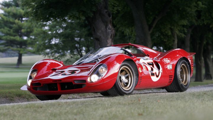 A picture is worth a thousand words ... - Page 1 - Ferrari Classics - PistonHeads - The image shows a vibrant red sports car parked on the grass near a road. It appears to be an old-fashioned race car, possibly from a vintage racing era. The car is adorned with a prominent number 69 and a logo that seems to represent a sponsor or team. It's positioned against the backdrop of a lush tree, adding a natural contrast to the man-made vehicle. The setting suggests this could be part of a race event or a vintage car showcase. The car exudes an air of nostalgia and speed.