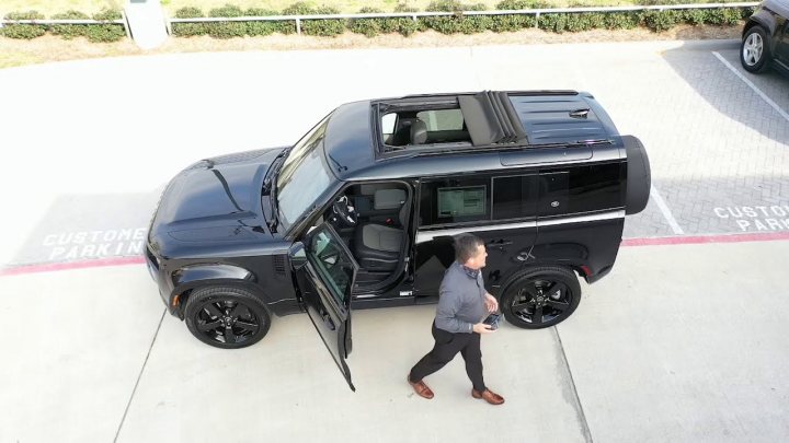 RE: Land Rover Defender Hard Top 110 | PH Review - Page 6 - General Gassing - PistonHeads UK - The image shows a black SUV parked in an indoor parking lot. A man is standing by the open door of the vehicle, seemingly about to enter or exit it. The interior of the car can be partially seen, with the driver's seat visible. There are no distinctive features on the SUV that suggest any modifications. The setting appears to be a garage with an outdoor parking space adjacent to a building facade. The man is dressed in casual attire and seems to be engaged in some activity related to the vehicle or his surroundings.