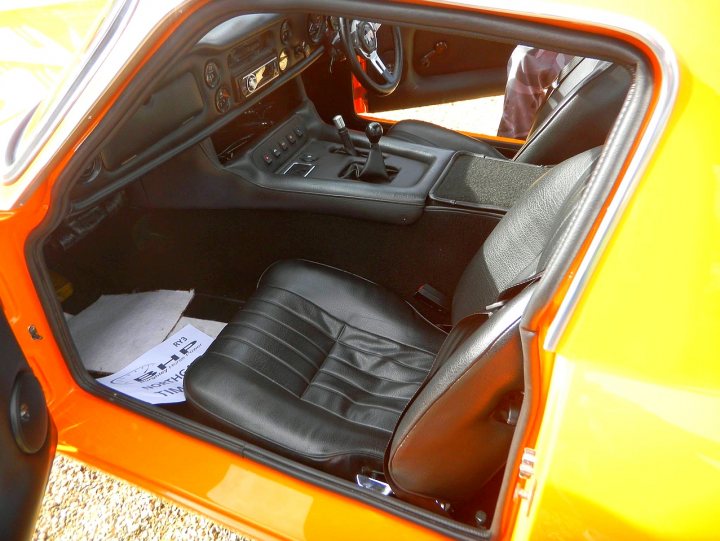 Early TVR Pictures - Page 90 - Classics - PistonHeads - The image captures the interior of a compact car. The perspective is from the outside looking into the driver's side. The interior is primarily black, with black seats and a black center console. A key feature of the dash is the shift knob, which adds a touch of orange color to the cabin. The floor mat, also in black, is lying in front of the console, slightly out of place, perhaps by the door or gear shift. The steering wheel, matching the overall color scheme, contributes to the overall harmony of the car's interior.